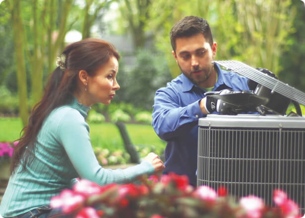 A man and woman look at an HVAC unit surrounded by flowers